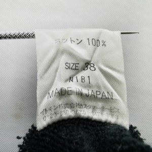 以下が含まれることがあります： 「コットン 100%」、「SIZE 38」、「N1B1」、および「MADE IN JAPAN」を含む黒いテキストの白いタグ。 タグは濃い色の生地に付いています。 銀色の針が挿入されています。