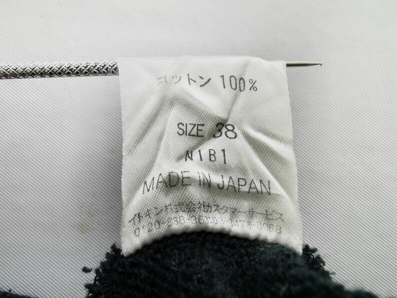 以下が含まれることがあります： 「コットン 100%」、「SIZE 38」、「N1B1」、および「MADE IN JAPAN」を含む黒いテキストの白いタグ。 タグは濃い色の生地に付いています。 銀色の針が挿入されています。