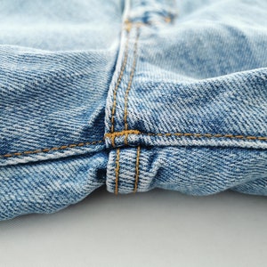 May include: Close-up of light blue denim jeans. The image highlights the details of the fabric, including the texture and stitching. The seams are sewn with a contrasting orange thread, adding visual interest.