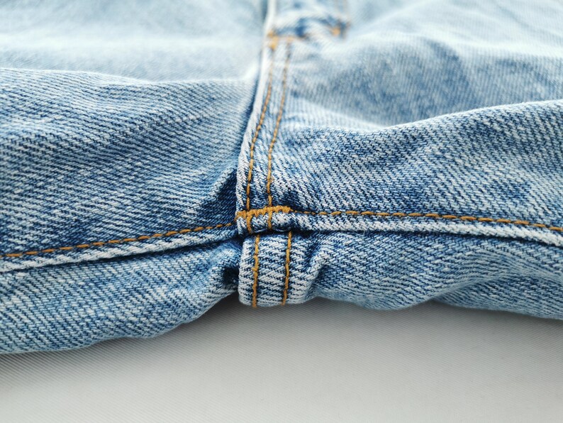 May include: Close-up of light blue denim jeans. The image highlights the details of the fabric, including the texture and stitching. The seams are sewn with a contrasting orange thread, adding visual interest.
