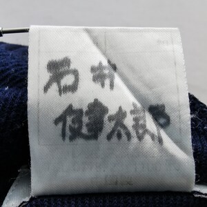 Puede incluir: Una etiqueta de tela blanca con caracteres japoneses impresos en tinta negra. La etiqueta est&aacute; adherida a una tela de punto azul oscuro. Una herramienta plateada sujeta la etiqueta.