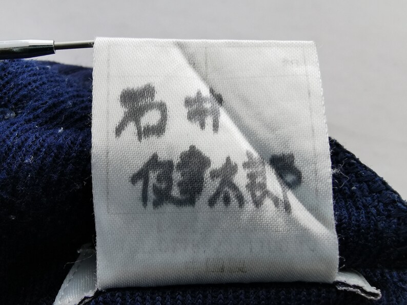 Puede incluir: Una etiqueta de tela blanca con caracteres japoneses impresos en tinta negra. La etiqueta est&aacute; adherida a una tela de punto azul oscuro. Una herramienta plateada sujeta la etiqueta.