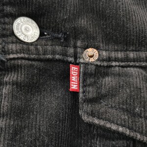 Puede incluir: Primer plano de una chaqueta de pana negra con un forro oscuro y difuso. La chaqueta presenta botones met&aacute;licos y una peque&ntilde;a etiqueta roja con la marca "EDWIN" en letras blancas. La pana tiene una textura acanalada.