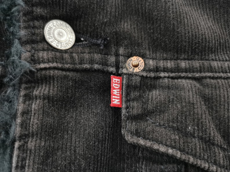 Puede incluir: Primer plano de una chaqueta de pana negra con un forro oscuro y difuso. La chaqueta presenta botones met&aacute;licos y una peque&ntilde;a etiqueta roja con la marca "EDWIN" en letras blancas. La pana tiene una textura acanalada.