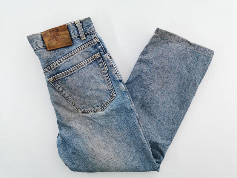 Puede incluir: Vaqueros de mezclilla azul claro con un dise&ntilde;o cl&aacute;sico de cinco bolsillos. Los vaqueros presentan un lavado descolorido y un parche de cuero en la cintura trasera. Los vaqueros est&aacute;n doblados y colocados sobre una superficie blanca.
