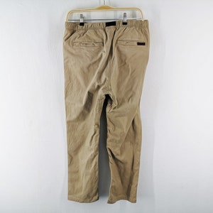 May include: Khaki-colored pants hanging on a wooden hanger against a white wall. The pants have two back pockets and a black label. The pants are made of a soft, durable material, and are suitable for casual wear.