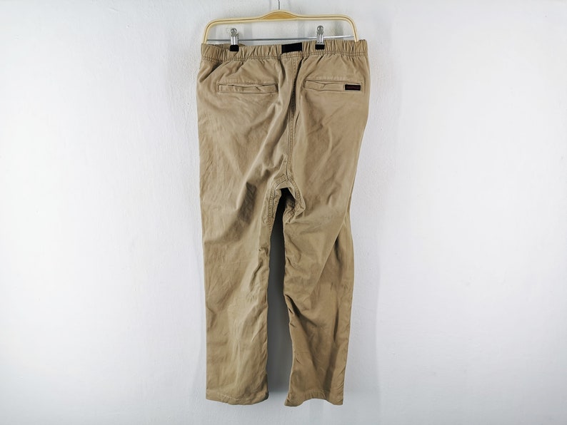 May include: Khaki-colored pants hanging on a wooden hanger against a white wall. The pants have two back pockets and a black label. The pants are made of a soft, durable material, and are suitable for casual wear.