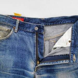 May include: Close-up of blue denim jeans with a partially unzipped fly. The jeans show signs of wear and fading, with a small pocket on the front and a button closure. A red tag with white text is visible.
