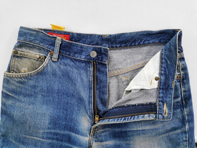 May include: Close-up of blue denim jeans with a partially unzipped fly. The jeans show signs of wear and fading, with a small pocket on the front and a button closure. A red tag with white text is visible.
