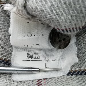 以下が含まれることがあります： 格子柄のグレーの生地のクローズアップ、日本語の文字が入った白いタグ、黒いボタン。タグにはサイズ「L」と記載されています。銀色のツールが前景にあります。