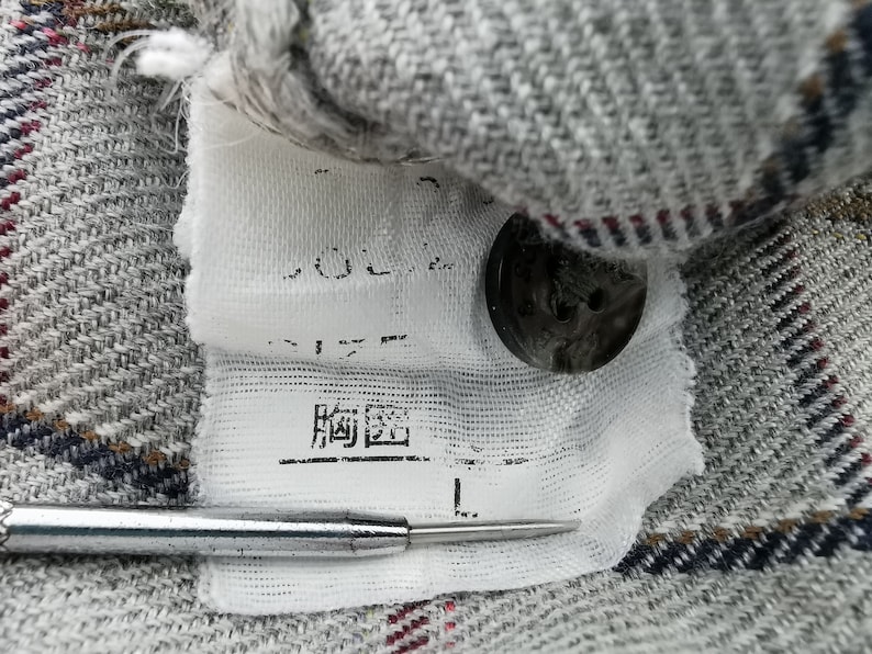 以下が含まれることがあります： 格子柄のグレーの生地のクローズアップ、日本語の文字が入った白いタグ、黒いボタン。タグにはサイズ「L」と記載されています。銀色のツールが前景にあります。