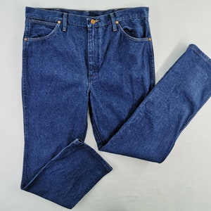 Puede incluir: Vaqueros de mezclilla azul oscuro con un dise&ntilde;o cl&aacute;sico de cinco bolsillos. Los vaqueros tienen cierre de bot&oacute;n y cremallera, junto con remaches de color cobre. Los pantalones est&aacute;n planos, mostrando el estilo de pierna recta.