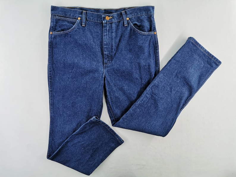 Puede incluir: Vaqueros de mezclilla azul oscuro con un dise&ntilde;o cl&aacute;sico de cinco bolsillos. Los vaqueros tienen cierre de bot&oacute;n y cremallera, junto con remaches de color cobre. Los pantalones est&aacute;n planos, mostrando el estilo de pierna recta.
