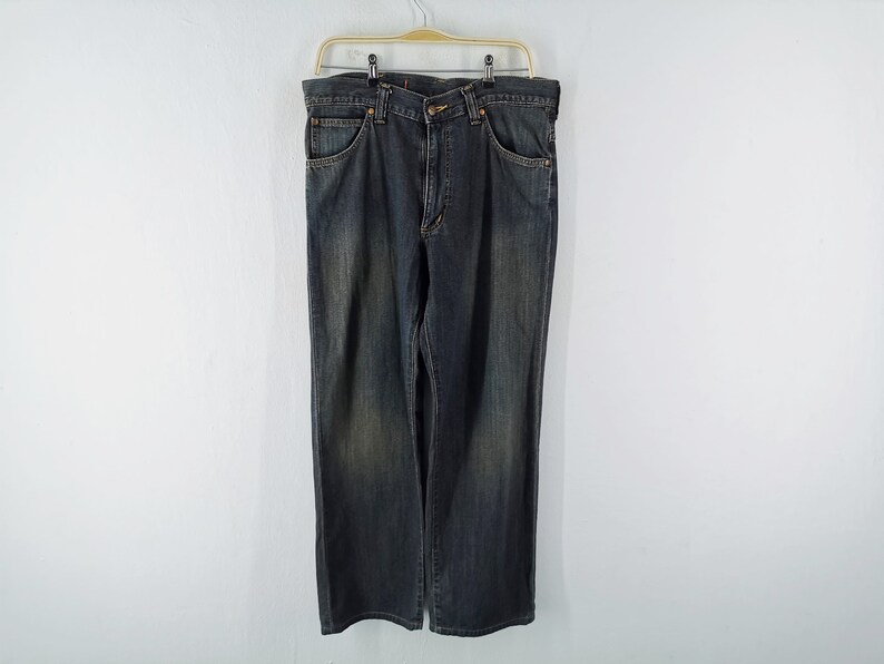 Peut inclure: Un jean en denim d&eacute;lav&eacute; fonc&eacute; suspendu &agrave; un cintre sur fond blanc. Le jean a une conception classique &agrave; cinq poches, une fermeture &agrave; bouton et &agrave; glissi&egrave;re, et une coupe droite. Le denim a un aspect d&eacute;lav&eacute; et us&eacute;.