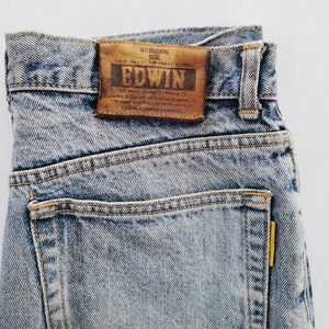 Op de afbeelding: Close-up van lichtblauwe denim jeans met een bruine leren patch. Op de patch staat "EDWIN" en andere tekst. De jeans heeft een klassiek ontwerp met vijf zakken, gele stiksels en een geel label op de achterzak.