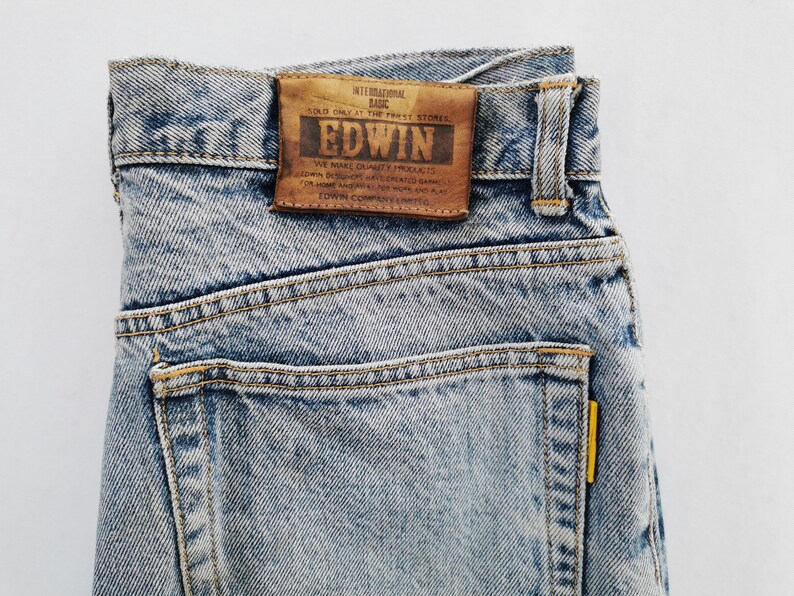 Op de afbeelding: Close-up van lichtblauwe denim jeans met een bruine leren patch. Op de patch staat "EDWIN" en andere tekst. De jeans heeft een klassiek ontwerp met vijf zakken, gele stiksels en een geel label op de achterzak.