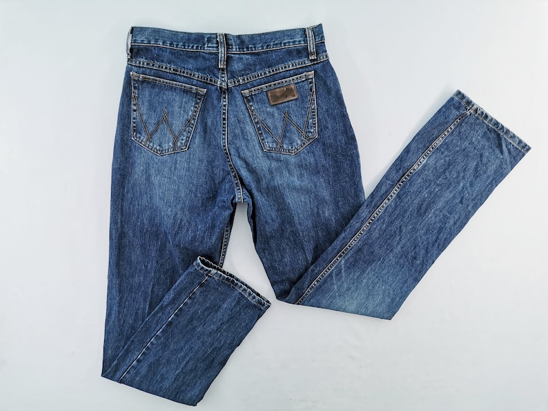 May include: Blue denim jeans with a classic five-pocket design. The jeans feature a brown leather patch on the back pocket with the brand logo. The jeans are laid flat on a white surface, showcasing the back pockets and the Wrangler logo.