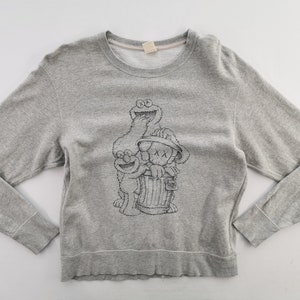 K&ouml;nnte beinhalten: Graues Sweatshirt mit Rundhalsausschnitt und langen &Auml;rmeln, verziert mit einer schwarz-wei&szlig;en Grafik von Sesame Street-Charakteren und einer KAWS-Figur. Die Grafik zeigt Kr&uuml;melmonster, Elmo und eine Figur in einer M&uuml;lltonne. Der Saum und die B&uuml;ndchen sind gerippt.
