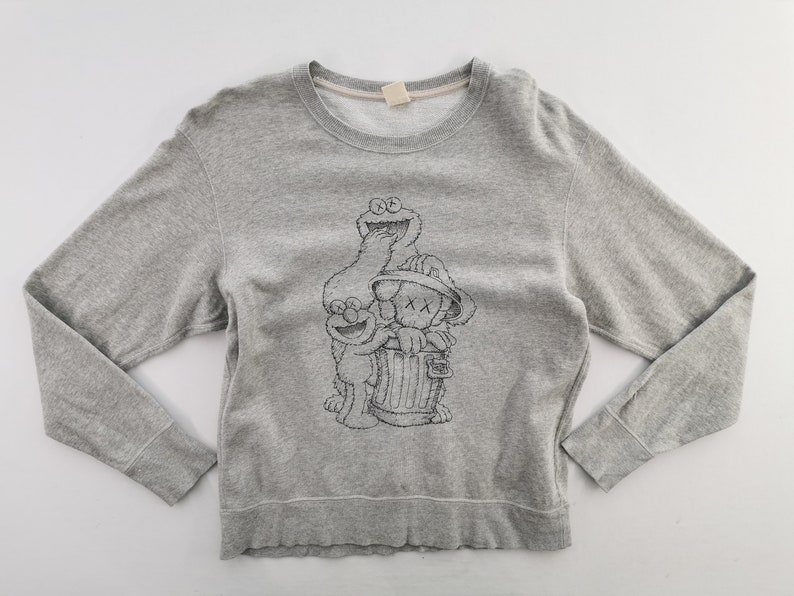 K&ouml;nnte beinhalten: Graues Sweatshirt mit Rundhalsausschnitt und langen &Auml;rmeln, verziert mit einer schwarz-wei&szlig;en Grafik von Sesame Street-Charakteren und einer KAWS-Figur. Die Grafik zeigt Kr&uuml;melmonster, Elmo und eine Figur in einer M&uuml;lltonne. Der Saum und die B&uuml;ndchen sind gerippt.