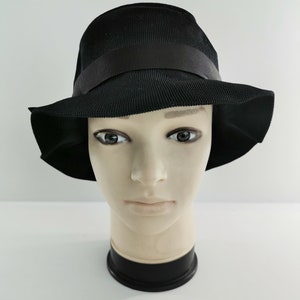 May include: A black hat with a wide brim and a black ribbon band. The hat is displayed on a mannequin head against a white background. The hat's material appears to be woven.