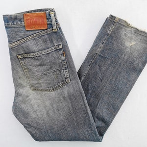 May include: Faded blue denim jeans with a classic five-pocket design. Features a leather patch with the brand name and size details. The jeans have a straight leg cut and show signs of wear.