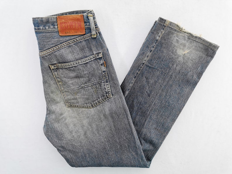 May include: Faded blue denim jeans with a classic five-pocket design. Features a leather patch with the brand name and size details. The jeans have a straight leg cut and show signs of wear.