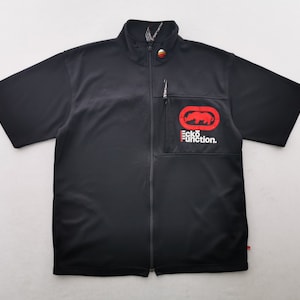 May include: A black short-sleeved zip-up shirt featuring a mock neck collar. The chest displays a red rhino logo inside an oval frame with the white text Ecko Function below it. A small red, orange, and yellow circular emblem sits at the collar.