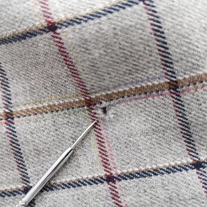 以下が含まれることがあります： ネイビー、ブラウン、レッド、イエローのチェック柄のグレーの生地のクローズアップ。銀色の針が小さな穴を指し、テキスタイルの詳細を強調しています。