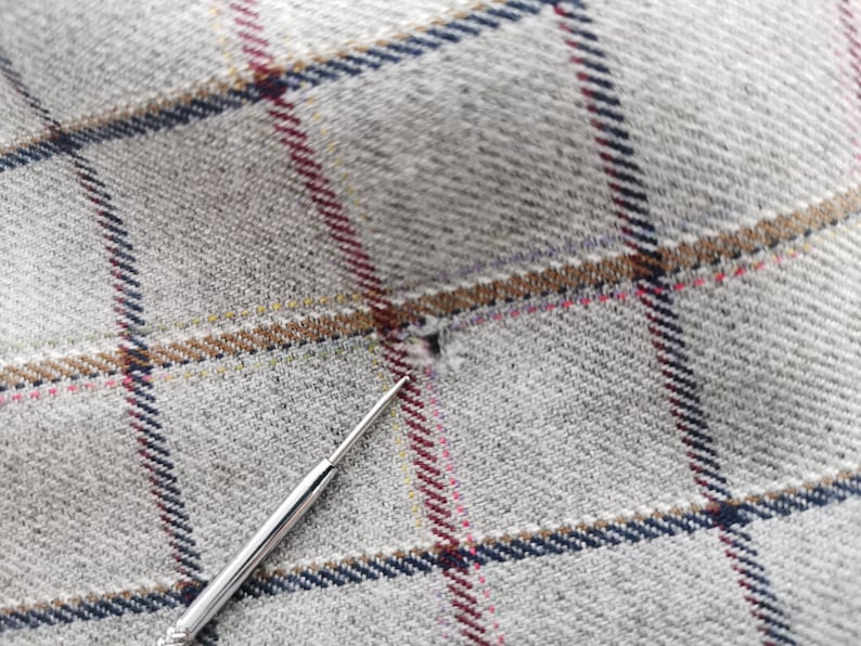 以下が含まれることがあります： ネイビー、ブラウン、レッド、イエローのチェック柄のグレーの生地のクローズアップ。銀色の針が小さな穴を指し、テキスタイルの詳細を強調しています。