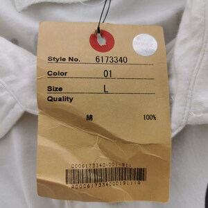 Puede incluir: Primer plano de una camisa blanca con una etiqueta adjunta. La etiqueta muestra el n&uacute;mero de estilo 6173340, el color 01 y la talla L. La etiqueta tambi&eacute;n indica que la camisa es 100% algod&oacute;n.
