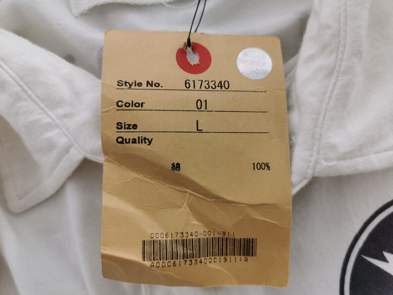 Puede incluir: Primer plano de una camisa blanca con una etiqueta adjunta. La etiqueta muestra el n&uacute;mero de estilo 6173340, el color 01 y la talla L. La etiqueta tambi&eacute;n indica que la camisa es 100% algod&oacute;n.