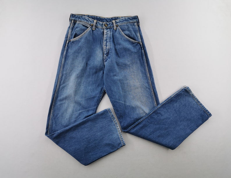 May include: Blue denim jeans with a button fly and classic five-pocket design. The jeans have a faded wash and a relaxed fit. The legs are slightly flared at the bottom. The jeans are laid flat on a white surface.
