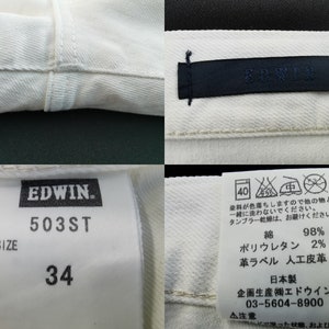 May include: Close-up of off-white denim jeans with visible stitching and tags. The tags display the brand "EDWIN", size 34, and care instructions. The fabric composition is 98% cotton and 2% polyurethane.