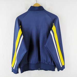 May include: A navy blue track jacket with a stand-up collar and a snap closure. The jacket has yellow and white stripes down the sleeves. The jacket is hanging on a wooden hanger against a white wall.