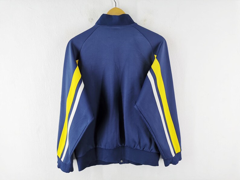 May include: A navy blue track jacket with a stand-up collar and a snap closure. The jacket has yellow and white stripes down the sleeves. The jacket is hanging on a wooden hanger against a white wall.