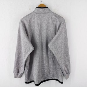 Puede incluir: Una sudadera gris de forro polar con ribete negro en el cuello y el dobladillo. La camisa de manga larga est&aacute; colgada en una percha de madera sobre un fondo blanco. Se ve la parte trasera de la camisa.