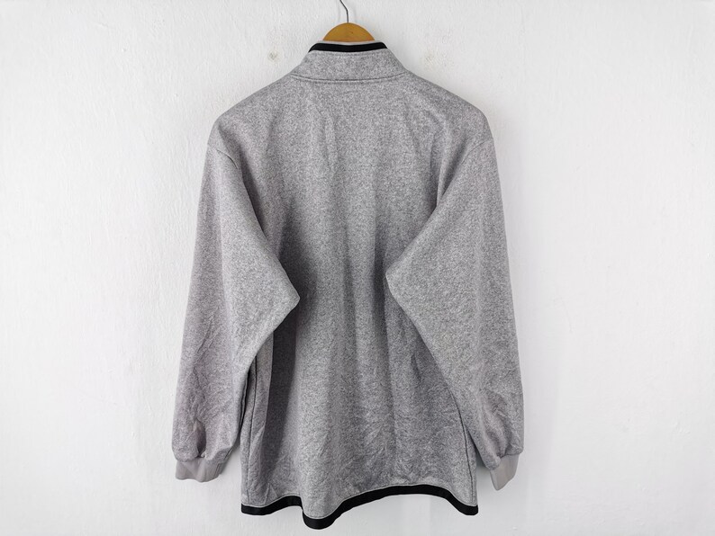 Puede incluir: Una sudadera gris de forro polar con ribete negro en el cuello y el dobladillo. La camisa de manga larga est&aacute; colgada en una percha de madera sobre un fondo blanco. Se ve la parte trasera de la camisa.