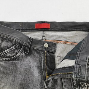 May include: Close-up of a pair of faded black denim jeans. The jeans have a button closure, a zipper, and pockets. A red tag is visible on the waistband. The jeans are displayed against a white background.