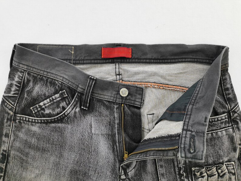 May include: Close-up of a pair of faded black denim jeans. The jeans have a button closure, a zipper, and pockets. A red tag is visible on the waistband. The jeans are displayed against a white background.