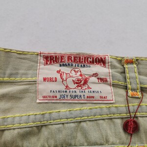 May include: Close-up of light tan denim jeans with yellow and orange stitching. A red and white True Religion brand label is visible, featuring a Buddha graphic and the text "WORLD TOUR".