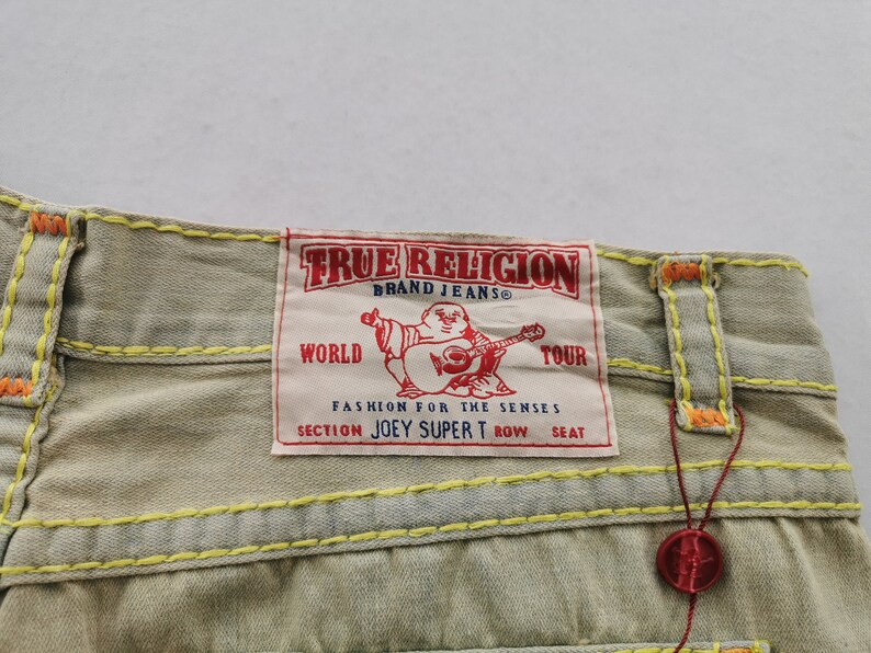 May include: Close-up of light tan denim jeans with yellow and orange stitching. A red and white True Religion brand label is visible, featuring a Buddha graphic and the text "WORLD TOUR".