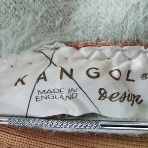 May include: Close-up of a white label with the brand name "KANGOL Design" and "MADE IN ENGLAND" printed in brown. The label is sewn onto a fuzzy, multi-colored fabric with shades of light blue, peach, and brown. A silver tool is in the foreground.