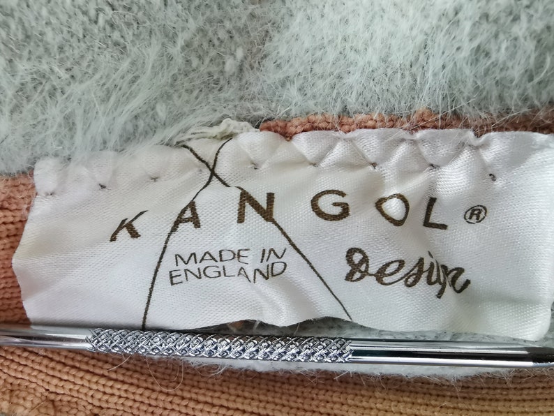 May include: Close-up of a white label with the brand name "KANGOL Design" and "MADE IN ENGLAND" printed in brown. The label is sewn onto a fuzzy, multi-colored fabric with shades of light blue, peach, and brown. A silver tool is in the foreground.