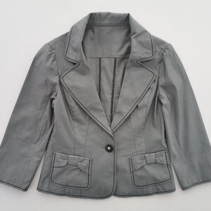 May include: A gray blazer with black stitching details. The blazer features a notched collar, a single button closure, and two front pockets with bow accents. The sleeves are three-quarter length.