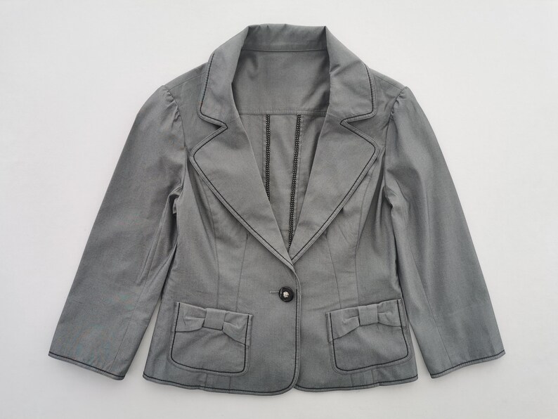 May include: A gray blazer with black stitching details. The blazer features a notched collar, a single button closure, and two front pockets with bow accents. The sleeves are three-quarter length.