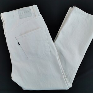 May include: Cream-coloured denim jeans with a classic five-pocket design. The jeans have a straight leg cut and a leather patch with the brand name. The back pocket has a black rivet. The jeans are folded on a black surface.