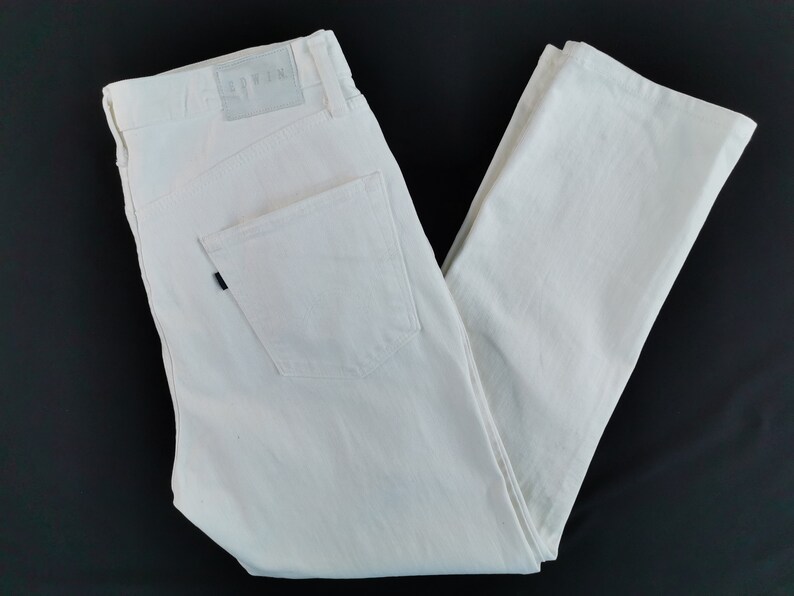 May include: Cream-coloured denim jeans with a classic five-pocket design. The jeans have a straight leg cut and a leather patch with the brand name. The back pocket has a black rivet. The jeans are folded on a black surface.