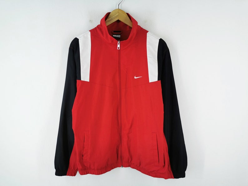 May include: Red, black, and white Nike track jacket with a full-length zipper. The jacket has a white Nike logo on the chest and white panels on the shoulders. The sleeves are black, and the body is red.