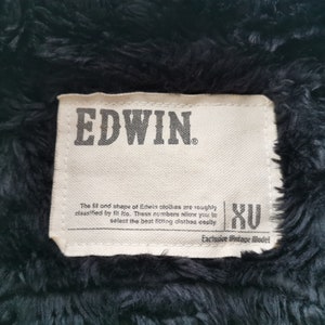 Puede incluir: Primer plano de una etiqueta de chaqueta Edwin. La etiqueta rectangular presenta el nombre de la marca "EDWIN" en letras grises oscuras. El texto describe el ajuste y la forma de la ropa Edwin, con la talla "XU". El fondo es una tela oscura y difusa.