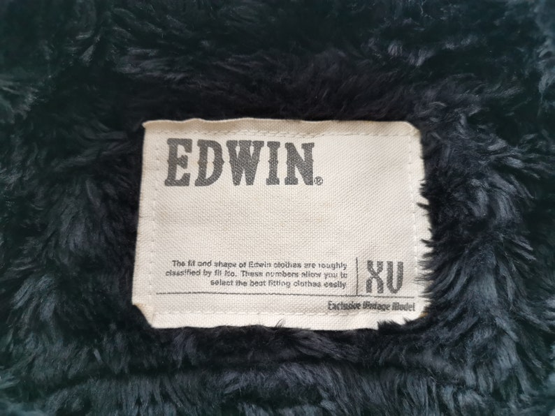 Puede incluir: Primer plano de una etiqueta de chaqueta Edwin. La etiqueta rectangular presenta el nombre de la marca "EDWIN" en letras grises oscuras. El texto describe el ajuste y la forma de la ropa Edwin, con la talla "XU". El fondo es una tela oscura y difusa.